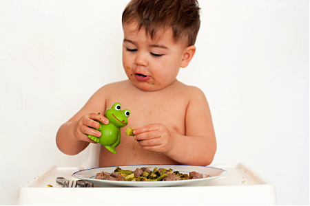 A dark-haired child in a high chair. There is a plate of food on the tray, and food on their hands and face. The child is feeding a green plastic frog from the plate.