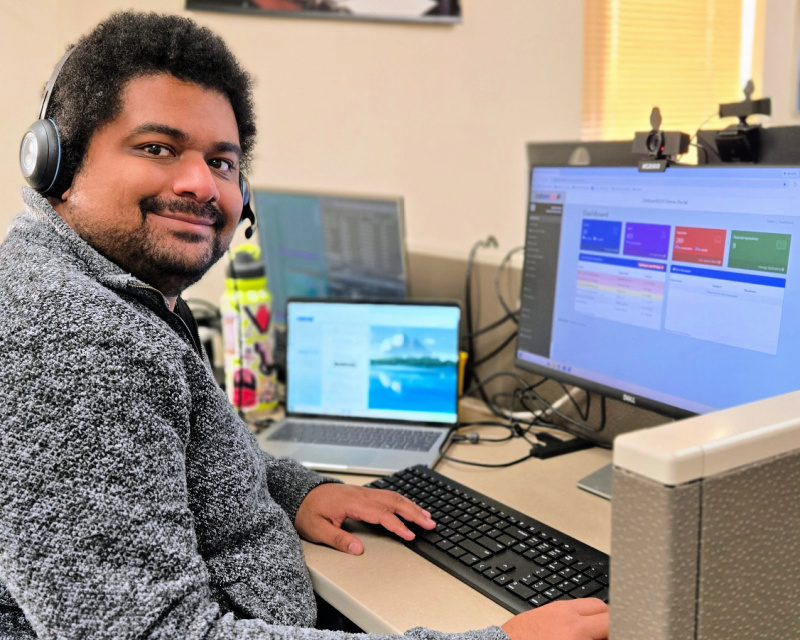 Andy Parris, a bearded man with short black hair, wearing a gray sweater, smiles in front of his desk at ClerkBase.