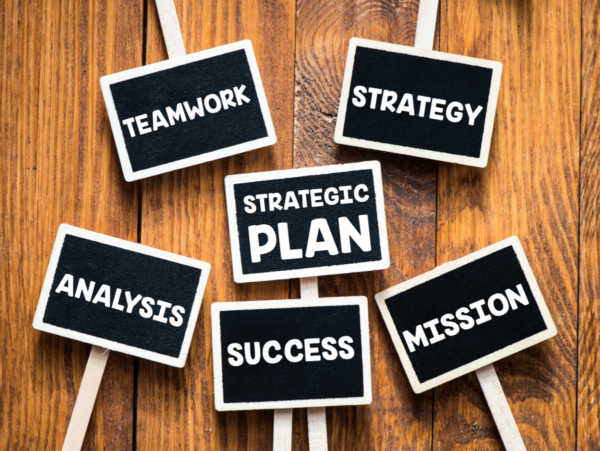 A collection of small stick signs on a wood plank background. The sign faces are black, with white lettering reading "teamwork," "strategy," "strategic plan," "analysis," "success," and "mission"